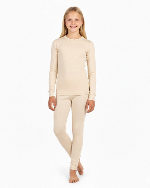 A girl wearing a natural cream-colored long-sleeve Merino wool base layer top and matching leggings, standing barefoot and smiling in a white studio.