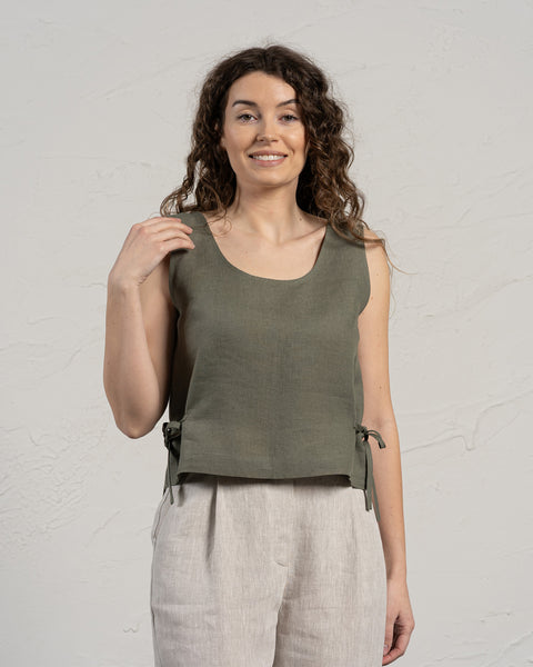 A smiling woman with long, curly brown hair wears a stone green sleeveless linen crop top with side ties and light-colored linen pants. She stands in front of a textured light gray wall, with one hand casually touching her shoulder.