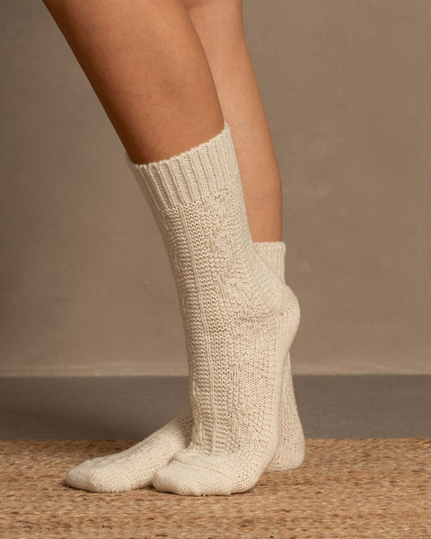 A low-angle shot shows a person's legs and feet, likely wearing menique brand off-white, crew-length knitted wool socks with a visible ribbed cuff and textured cable-knit pattern on the foot. The person is standing on a light brown, woven mat, against a neutral, plain background.