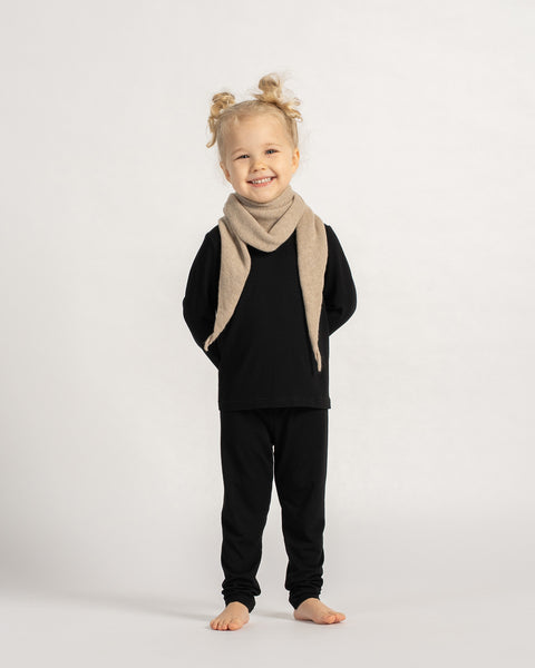 Little girl wearing a beige knit triangle scarf around her neck, dressed in a black outfit and smiling while standing barefoot against a neutral background.