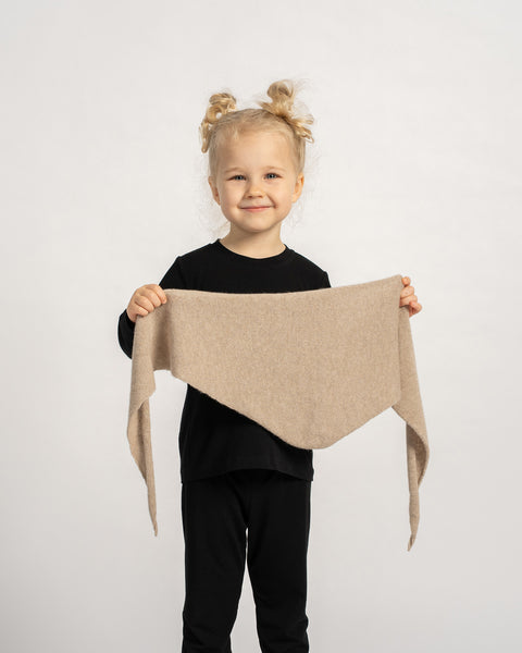Smiling child in a black outfit holding a beige knit triangle scarf, showing its shape and soft texture against a light background.