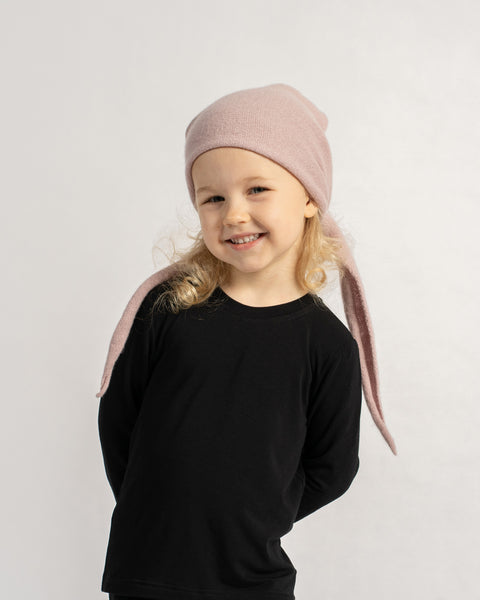 Smiling little girl in a black outfit wearing a pink knit headscarf styled as a hat with long ties hanging down.