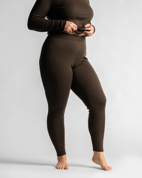 A barefoot woman posed at a three-quarter angle against a white background, showcasing the fit of a dark brown base layer leggings set.