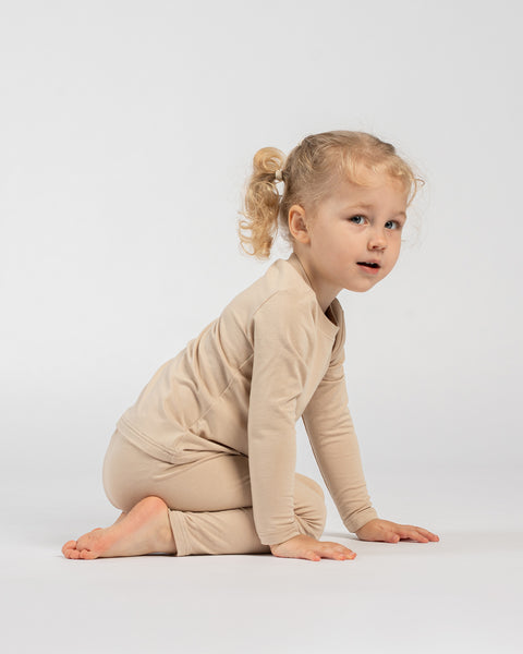 Child in beige long-sleeve outfit kneeling on the floor with hands resting in front, looking slightly to the side against a plain background.
