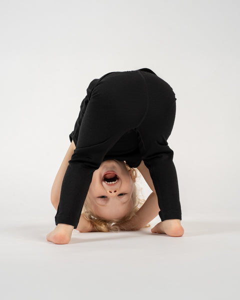 A young child with light-colored hair is playfully bent over with their head inverted and looking between their legs, directly at the viewer. They are wearing dark-colored pants and their bare feet are visible. The child has a wide, open-mouthed smile, and their expression is joyful. The background is a plain white surface.