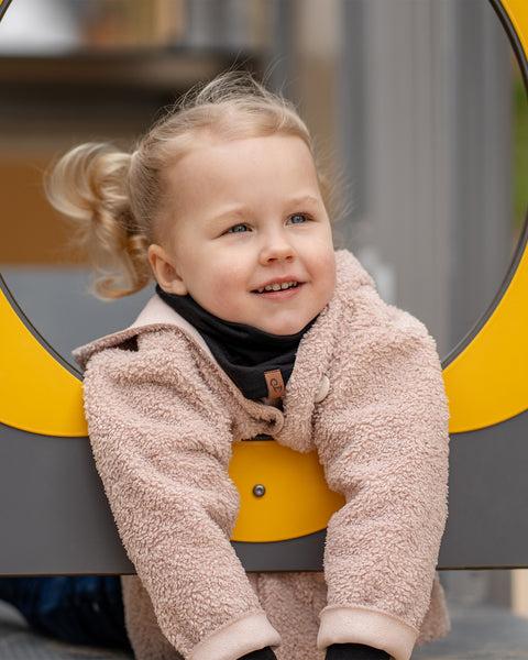 A cheerful child with blonde pigtails looks upwards and to the right, peering through a yellow and gray circular opening on a playground. The child is wearing a dark Menique neck gaiter and a fluffy beige jacket, with a slight smile on their face.