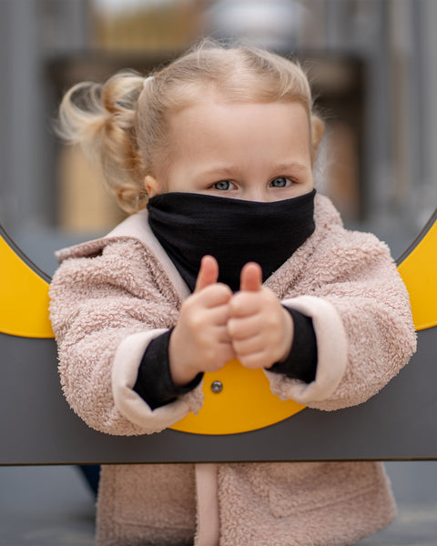 A cheerful child with blonde pigtails looks straight and to the left, peering through a yellow and gray circular opening on a playground. The child is wearing a Black Menique neck gaiter and a fluffy beige jacket, with a slight smile on their face.