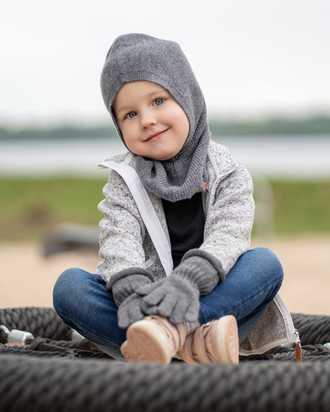 A young child with blue eyes and a slight smile, sits cross-legged on a black rope swing, looking directly at the viewer. They are wearing a dark grey balaclava, a grey speckled zip-up hoodie, dark grey mittens, blue jeans, and light pink sneakers. The background is a blurred outdoor scene with sand, grass, and water.