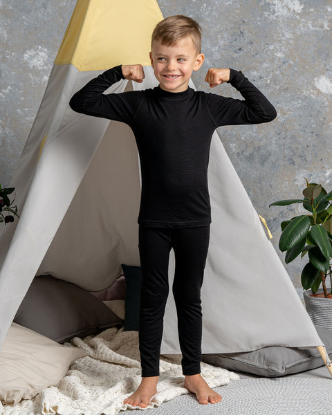 a boy wearing the Kids' 160 Merino Pants in Black color. He has his arms raised up, acting strong. Behind him is a children's indoor play tent.