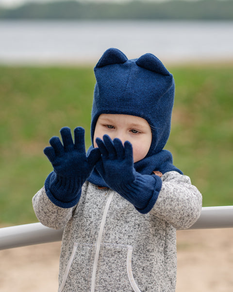 A young child wearing a menique dark blue balaclava with ears, dark blue gloves, and a grey speckled zip-up hoodie, holds their hands up in front of their face, looking slightly downward. The background is a blurred outdoor scene with green grass and a body of water.
