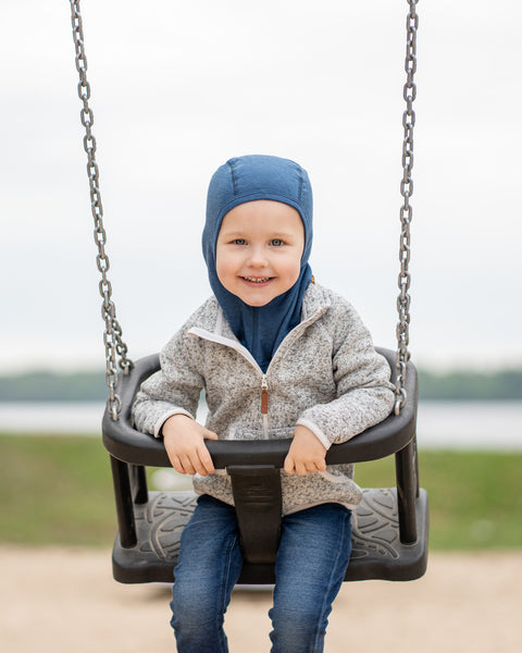 A young child with blonde hair and a happy expression, sits in a black swing on a playground. They are wearing a menique denim color balaclava, a grey speckled zip-up hoodie, and blue jeans, looking directly at the camera with a blurred background of water and grass.