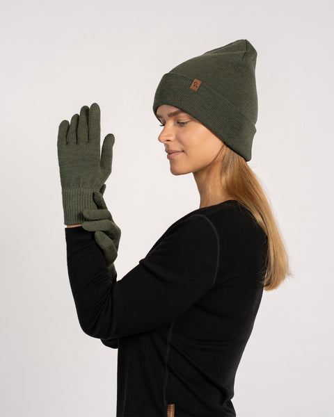 Woman wearing a dark green knit beanie and matching gloves, dressed in a black long-sleeve top, shown in profile view with one gloved hand raised against a plain background.