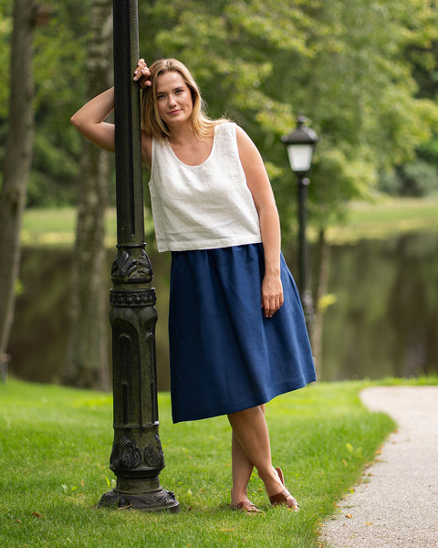 A woman leans comfortably against an ornate black lamppost in a grassy park setting next to a tranquil lake. She is wearing a pure white linen tank top, a storm blue midi skirt, and brown leather sandals.
