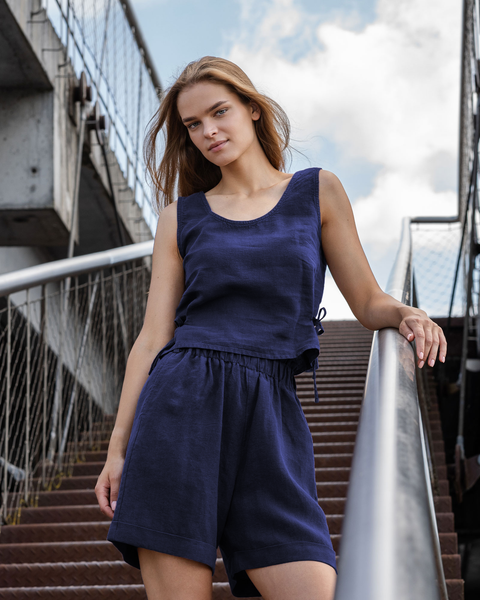 A full-body shot of a young woman with long, light brown hair standing on a metal staircase outdoors. She is looking directly at the camera with a neutral expression. She is wearing a two-piece linen outfit in a storm blue color, consisting of a sleeveless top with a scoop neck and high-waisted shorts with an elastic waistband. Her left arm is resting on the metal handrail beside her. The staircase has textured, possibly rusted, steps.