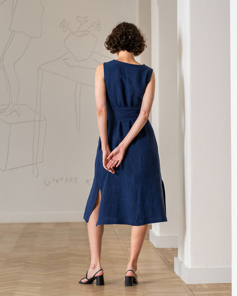 A woman with short, curly dark hair stands with her back to the viewer in a storm blue linen sleeveless dress and black heeled sandals. The dress has a slightly cinched waist with a matching fabric belt. Her hands are clasped gently in front of her. She is standing on a light-colored wooden floor in a room with a white wall featuring a faint, sketched design of figures and objects.