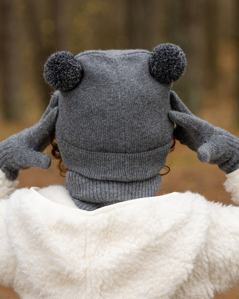 The back of a child's head and shoulders are shown, with the child wearing a dark grey knit hat with two dark grey pom-poms on top. They are also wearing a grey turtleneck and a cream-colored sherpa jacket, with dark grey mittens on their hands, against a blurred forest background.
