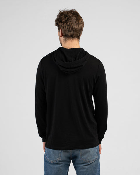 A studio shot showing the back of a man wearing a black long-sleeved hooded shirt with the hood down and blue jeans. He stands facing away from the camera against a plain light grey background.