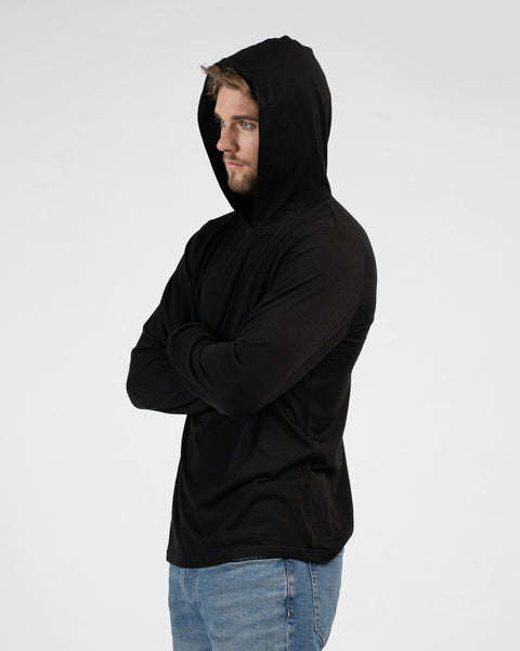 A side-profile studio portrait of a man looking off to the left. He stands with his arms crossed, wearing a black long-sleeved hooded shirt with the hood up and blue jeans.