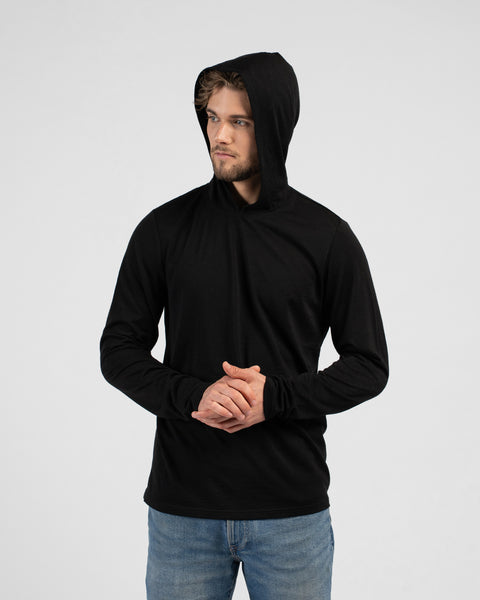 A studio portrait of a man looking off to the left side of the frame with his hands clasped in front of him. He is wearing a black long-sleeved hooded shirt with the hood pulled up and blue jeans.