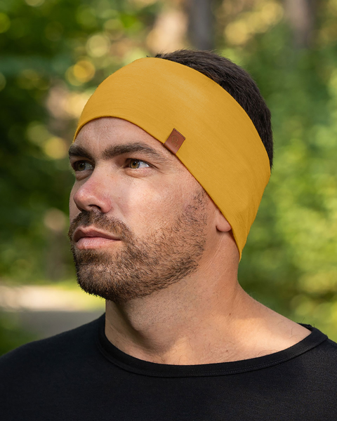 A man with short, dark hair and a beard is shown from the chest up, wearing a spicy yellow merino wool headband with a small brown tag. He is wearing a black shirt and looking off to his left with a neutral expression. The background is softly blurred, showing green foliage and tree trunks, suggesting an outdoor setting.