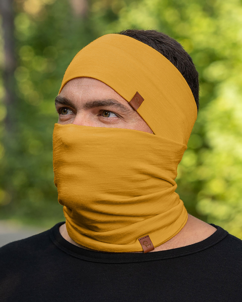 A person with short, dark hair is shown wearing a spicy yellow headband and a matching neck gaiter that is pulled up to cover their mouth and nose, with only their eyes visible. They are wearing a black long-sleeved shirt, and the blurred background suggests an outdoor setting with green foliage.