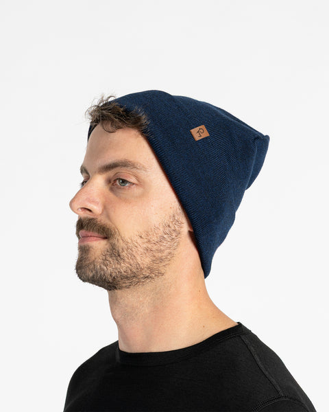 A person with light skin, short brown hair, and a beard is shown in profile against a white background. They are wearing a blue knit beanie with a small brown tag on the side. They are also wearing a black t-shirt.