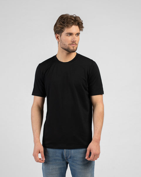 A studio portrait of a man looking off to the right side of the frame with his hands relaxed by his sides. He is wearing a black short-sleeved t-shirt and blue jeans.