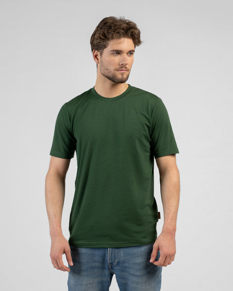 A studio portrait of a man standing against a plain white background. He is looking off to the right side of the frame. He has medium-length brown hair and a light beard. He is wearing a short-sleeved, dark green crew-neck t-shirt with a regular fit, paired with medium-wash blue jeans. His arms are relaxed by his sides. There is a small tag visible on the lower left hem of the t-shirt.