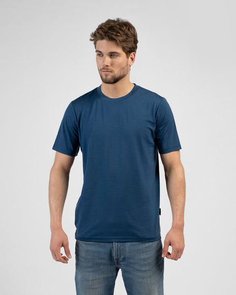 A studio portrait of a man standing against a plain white background. He is looking off to the left side of the frame with a neutral expression. He is wearing a short-sleeved, denim-blue crew-neck t-shirt with a regular fit, paired with medium-wash blue jeans. His arms are relaxed by his sides.