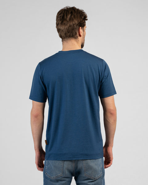 A studio shot showing the back of a man wearing a denim blue short-sleeved t-shirt and blue jeans. He stands facing away from the camera against a plain light grey background.