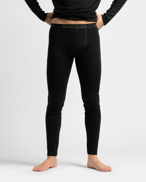 A model from the waist down wearing black men's compression leggings with a black waistband featuring the brand name 'MENIQUE' in gold lettering. The model is standing barefoot against a light gray studio background.