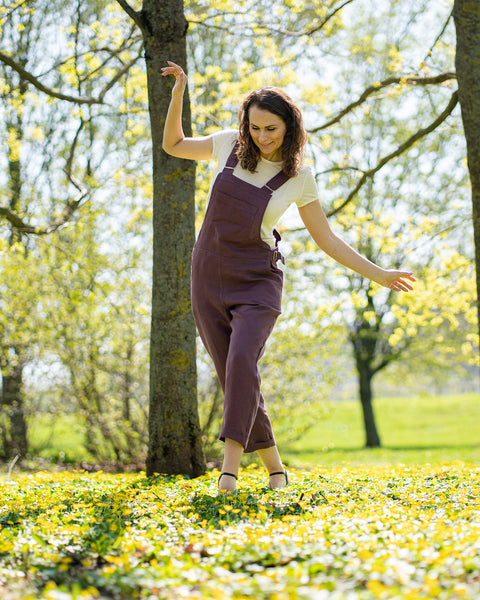 A woman with brown hair, wearing dark purple overalls over a light-colored t-shirt, stands outdoors between two tree trunks on grassy ground.