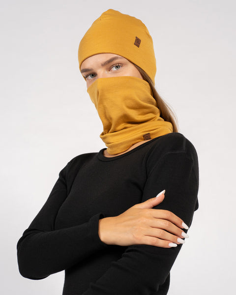 Model in black top styled with mustard yellow Merino beanie and neck gaiter covering face, arms crossed.