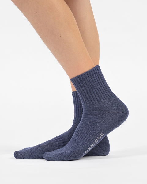 A side-profile view of a person's feet wearing heathered blue crew socks, revealing the word "MENIQUE" printed on the sole of the right foot.