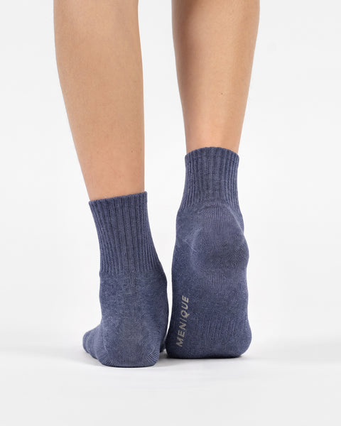 A back view of a person's lower legs and feet wearing heathered blue crew socks, with the right heel slightly lifted to show the "MENIQUE" logo on the sole.