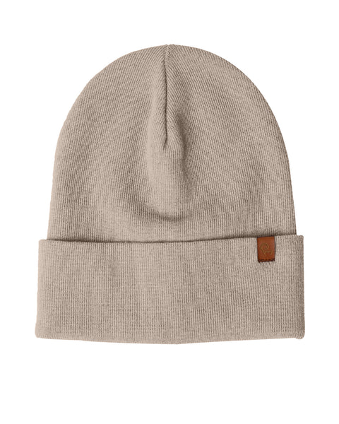 A creamy beige knit beanie is displayed against a white background. The beanie has a folded brim and a small brown rectangular tag is attached to the brim on the right side. The knit appears to be a fine gauge.