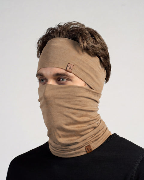 A man looking slightly to the left, wearing a matching beige headband and a neck gaiter pulled up over his nose and mouth, covering the lower half of his face. He has light eyes and brown hair. He is wearing a black long-sleeved top. Both the headband and the gaiter feature a small, rectangular brown leather logo tag. The background is a solid light grey.