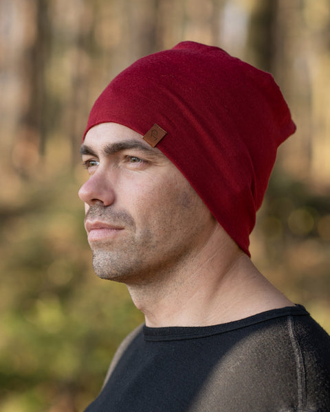 A man standing outside in a forest during fall season, wearing the menique Men's Merino Beanie Hat in Royal Cherry Color.