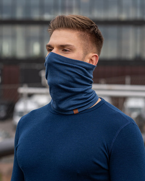 A man wearing the menique unisex denim neck gaiter. He is outside, wearing a matching color long sleeve merino top.