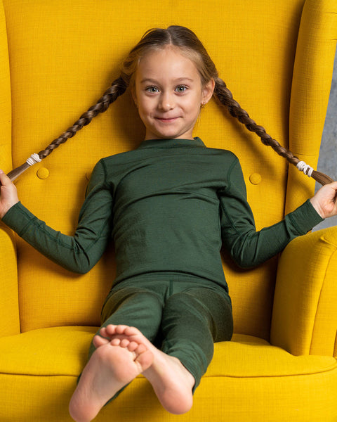 A girl smiling at the camera, wearing the menique Kids' Long Sleeve Crew 160 Merino in Dark Green color. She is sitting in a yellow ottoman, holding her braids with her hands to the sides.