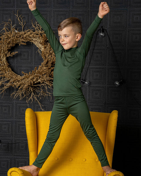 A boy wearing the menique kids' 160gsm merino set in dark green color. It ncludes a long sleev etop and pants. He has his arms raised up straight and legs wide apart, standing on a spicy yellow color ottoman.