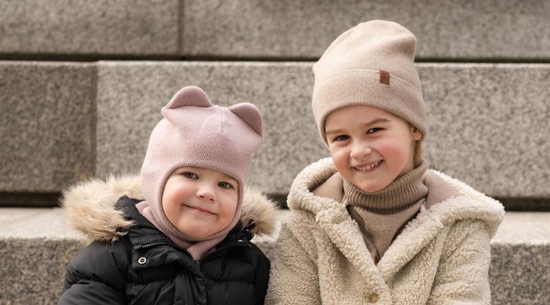 two girls sitting outside on stairs, wearing the menique knit kids' accessories (knit balaclava with ears in dusty pink and creamy beige knit beanie).