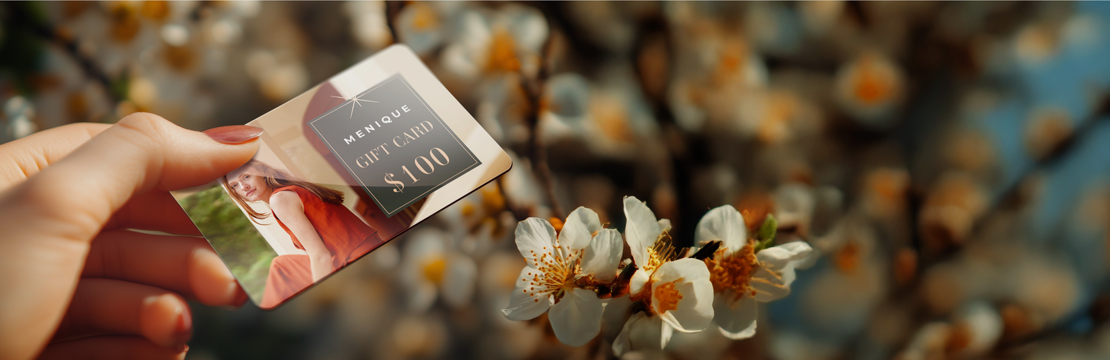 A wide, banner-style image showing a hand holding up a Menique gift card in the foreground. The gift card features an image of a woman in a red top and displays the text "MENIQUE GIFT CARD $100" in a dark grey box with gold lettering. The background is a beautifully blurred, shallow depth-of-field shot of white spring blossoms on a tree branch, bathed in warm sunlight.