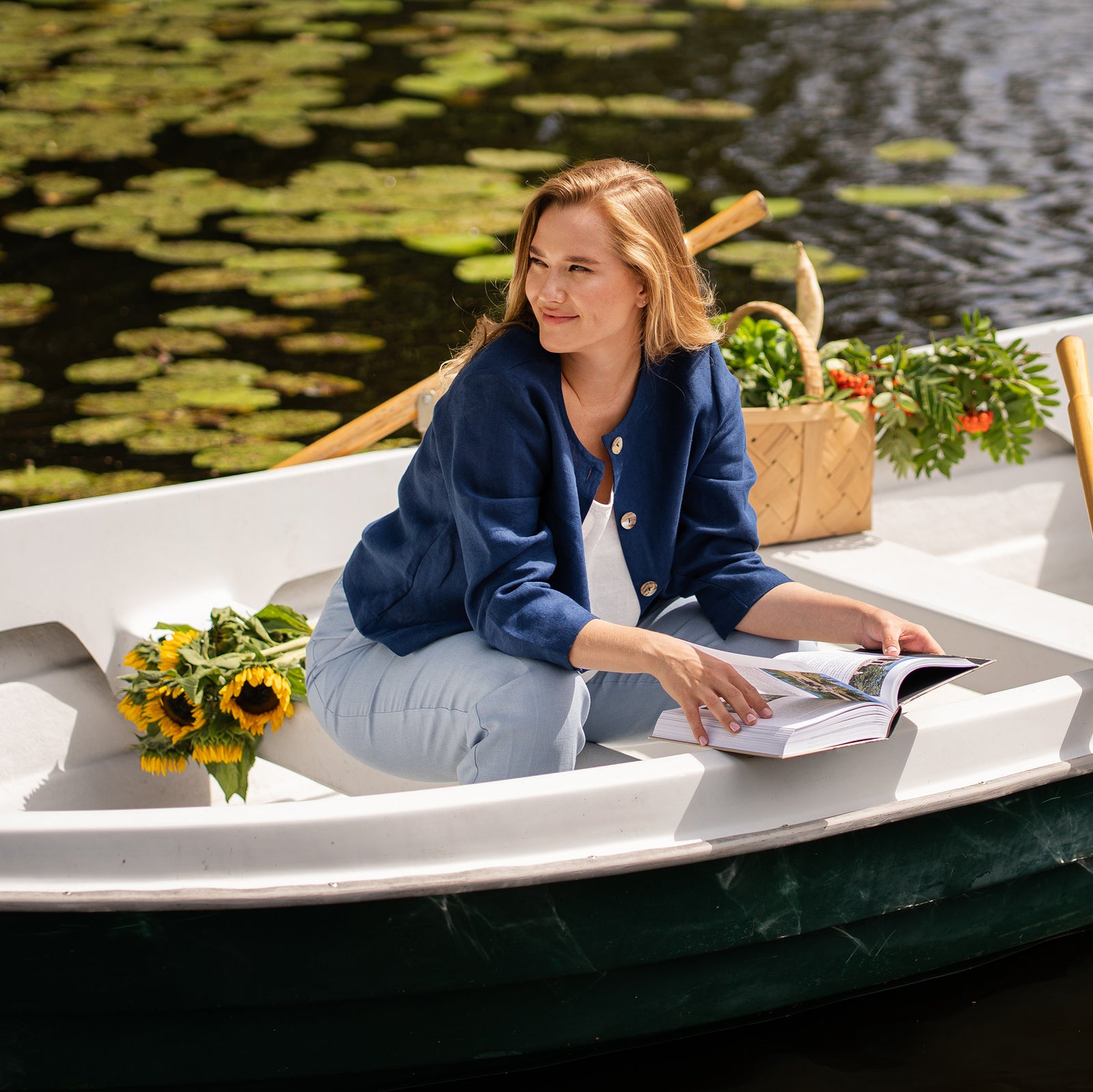 A woman reading a book in a rowboat, styled in a navy blue linen button-up jacket paired with light blue trousers.