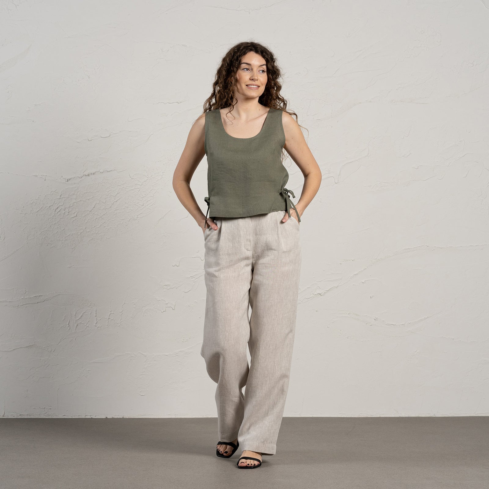 A smiling woman with long, curly brown hair wears a sleeveless linen crop top with side ties and light-colored wide-leg linen pants. She stands in front of a textured light gray wall, with one hand in her pocket.