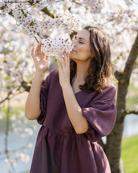 A smiling woman with dark, wavy hair is captured in a close-up, outdoor shot, gently bringing a cluster of white cherry blossoms to her nose to smell them. She has her eyes closed and appears to be enjoying the fragrance. She is wearing a shadow purple colored linen dress with a V-neck and elbow-length, puffed sleeves. The background features more cherry blossoms and a body of water.