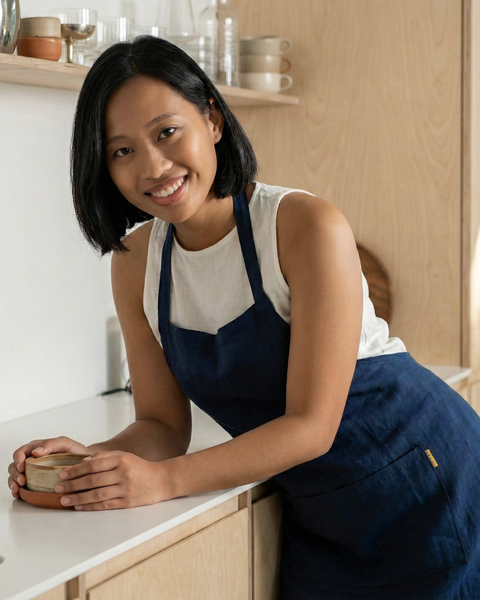 Smiling woman in a storm blue linen bib apron leaning on a kitchen counter, minimalist home interior.