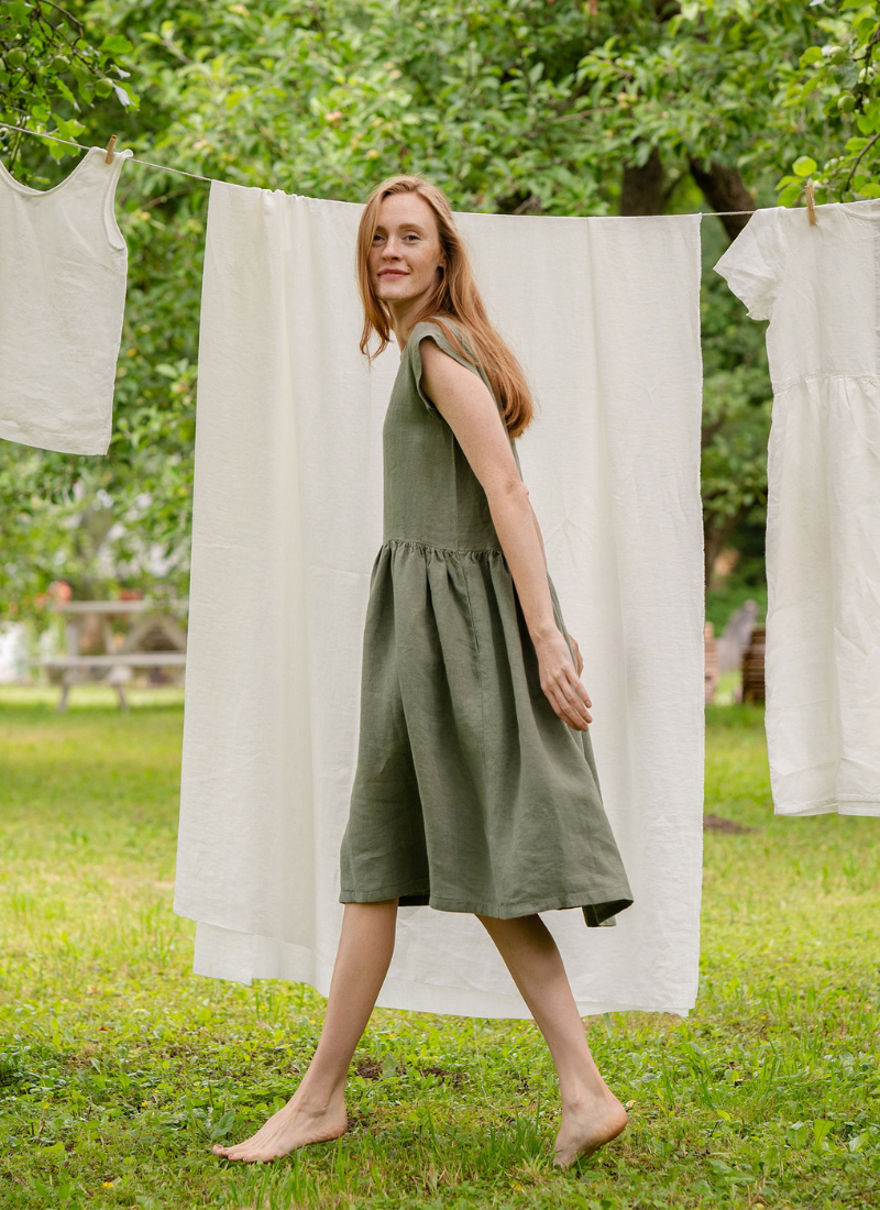 A barefoot woman wearing a sleeveless olive green linen dress is walking across a grassy garden. Behind her, light-colored linen garments are hanging on a clothesline between trees, creating a relaxed, sustainable lifestyle atmosphere. The natural setting emphasizes the breathable texture and effortless movement of the dress.