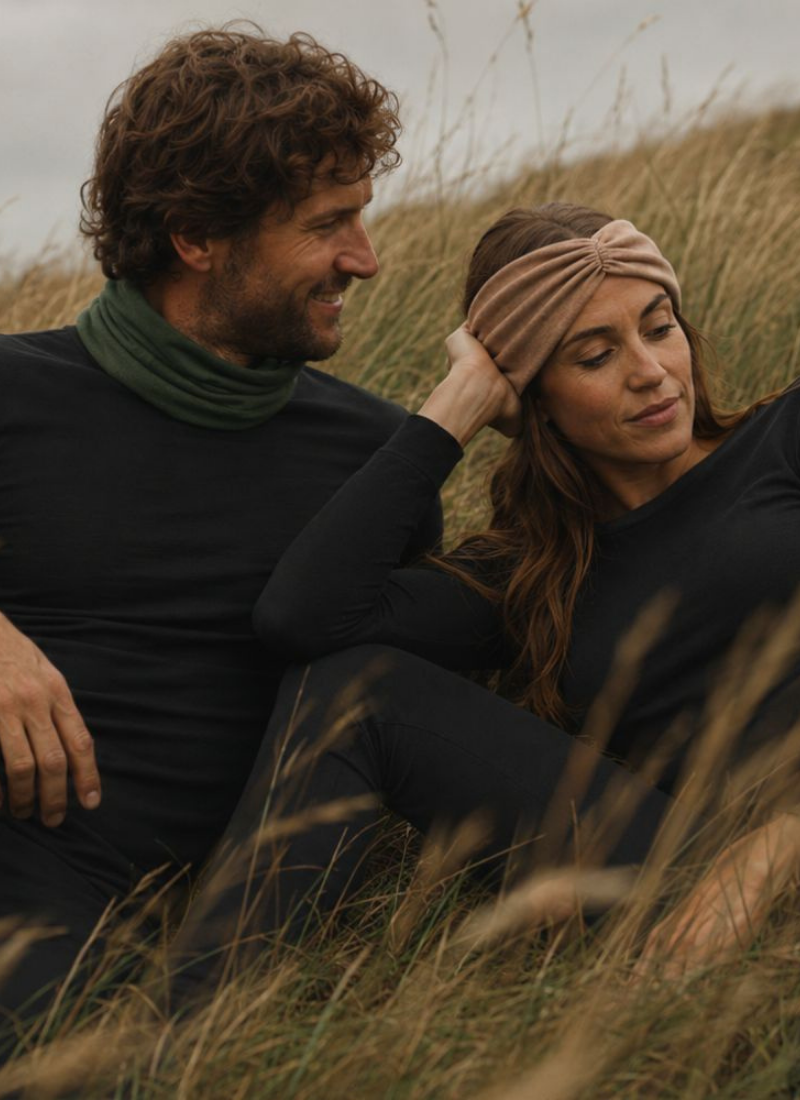 A smiling man and woman sit together in a field of tall, dry grass. The man wears a black long-sleeved top with a green neck gaiter, while the woman wears a matching black outfit accented by a tan twisted headband.