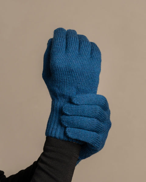 
A close-up shot of a person's hands wearing menique brand vibrant blue, ribbed knit gloves. The left hand is clenched into a fist, while the right hand appears to be gripping the wrist area of the left glove. The person is wearing a dark long-sleeved shirt underneath. The background is a solid, neutral gray.